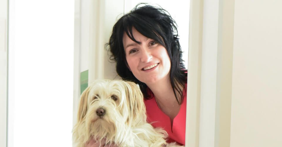 La Dottoressa Sara Coan della Clinica Veterinaria Serenissima a Sacile, in provincia di Pordenone, con i capelli scuri e un top rosa sorride tenendo in braccio un piccolo e soffice cane di colore chiaro in un ambiente interno molto illuminato, irradiando calore e spirito di squadra.