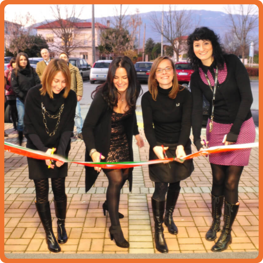 storia_mobile Le quattro socie fondatrici della Clinica Veterinaria Serenissima sorridono e tagliano con le forbici un nastro rosso e bianco all'aperto, con spettatori e auto parcheggiate sullo sfondo. Durante l’inaugurazione della clinica veterinaria a Sacile.